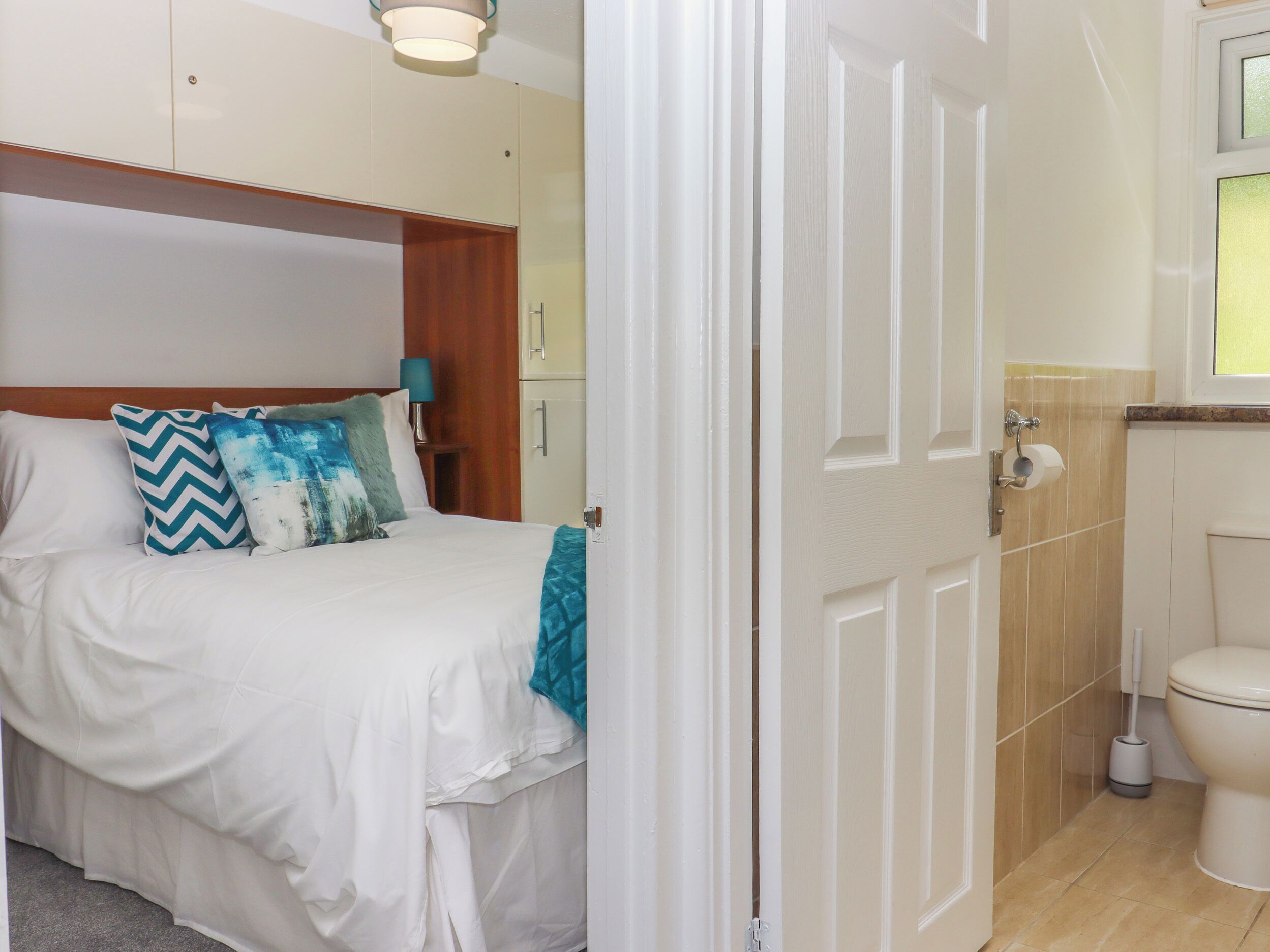 Quarto com cama, almofadas, mesa de cabeceira e porta que dá para um banheiro.