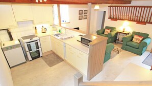 A compact kitchen with a microwave, oven, and sink. There is a green sofa and a small table with a lamp. - Y Stabl (Llanbedr)