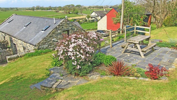 A stone building with a sloped roof, a red house, and a wooden bench in a garden with flowering plants. - Y Stabl (Llanbedr)
