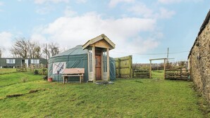 Een klein, rustiek houten gebouw met een schuin dak, mogelijk een tuinhuis of een kleine hut, in een grasrijke omgeving met een stenen muur en een metalen hek op de achtergrond.