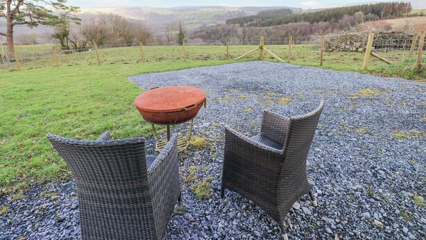 Een terras met rieten stoelen en een ronde tafel op een grindpad, omgeven door een grasveld en een hek, met uitzicht op verre heuvels.