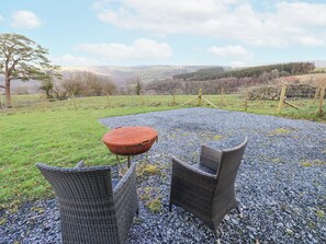 An outdoor seating area with wicker chairs and a round table on a gravel surface, surrounded by a grassy field and a fence, with a view of distant hills. - Blaidd Bach (Carmarthen)
