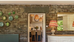 A dining area with a stone wall, a kitchen in the background, and a view of a garden through a window. - Leathes Head Country House (Keswick)