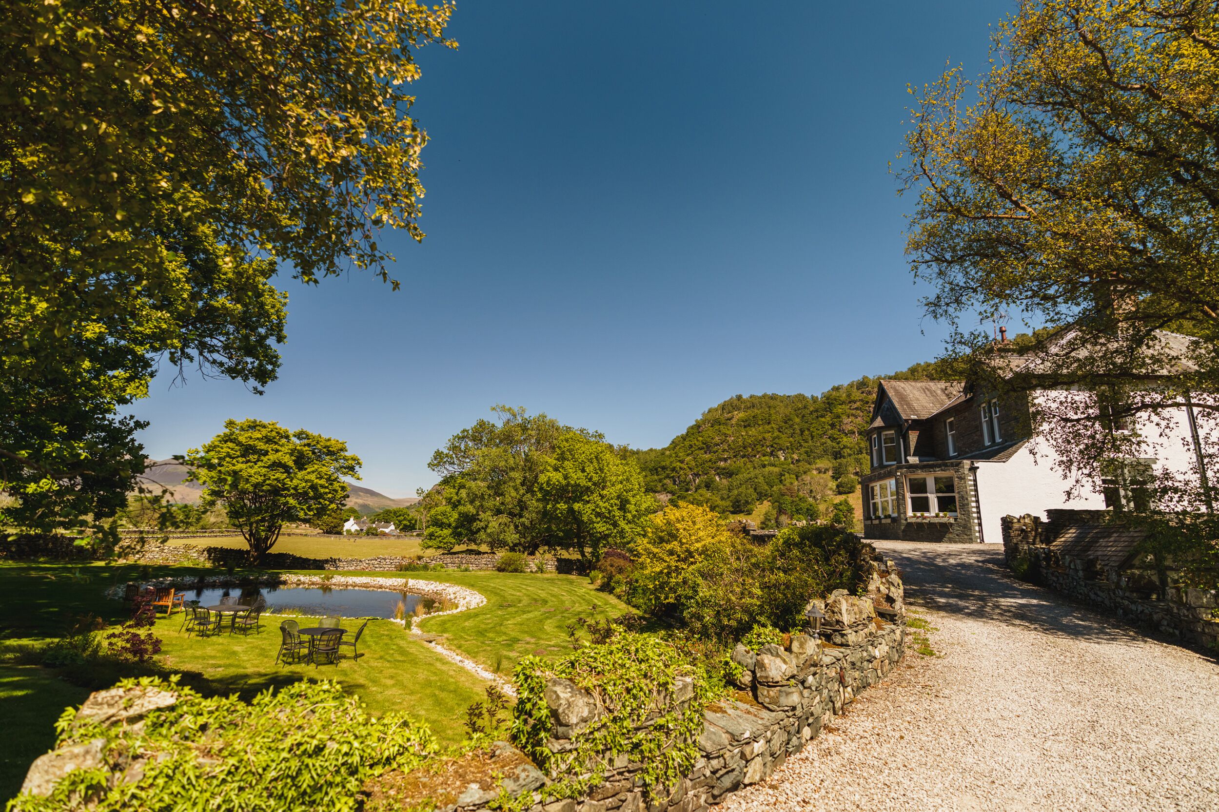 A house with a stone wall, a gravel driveway, and a lawn with a pond and chairs.