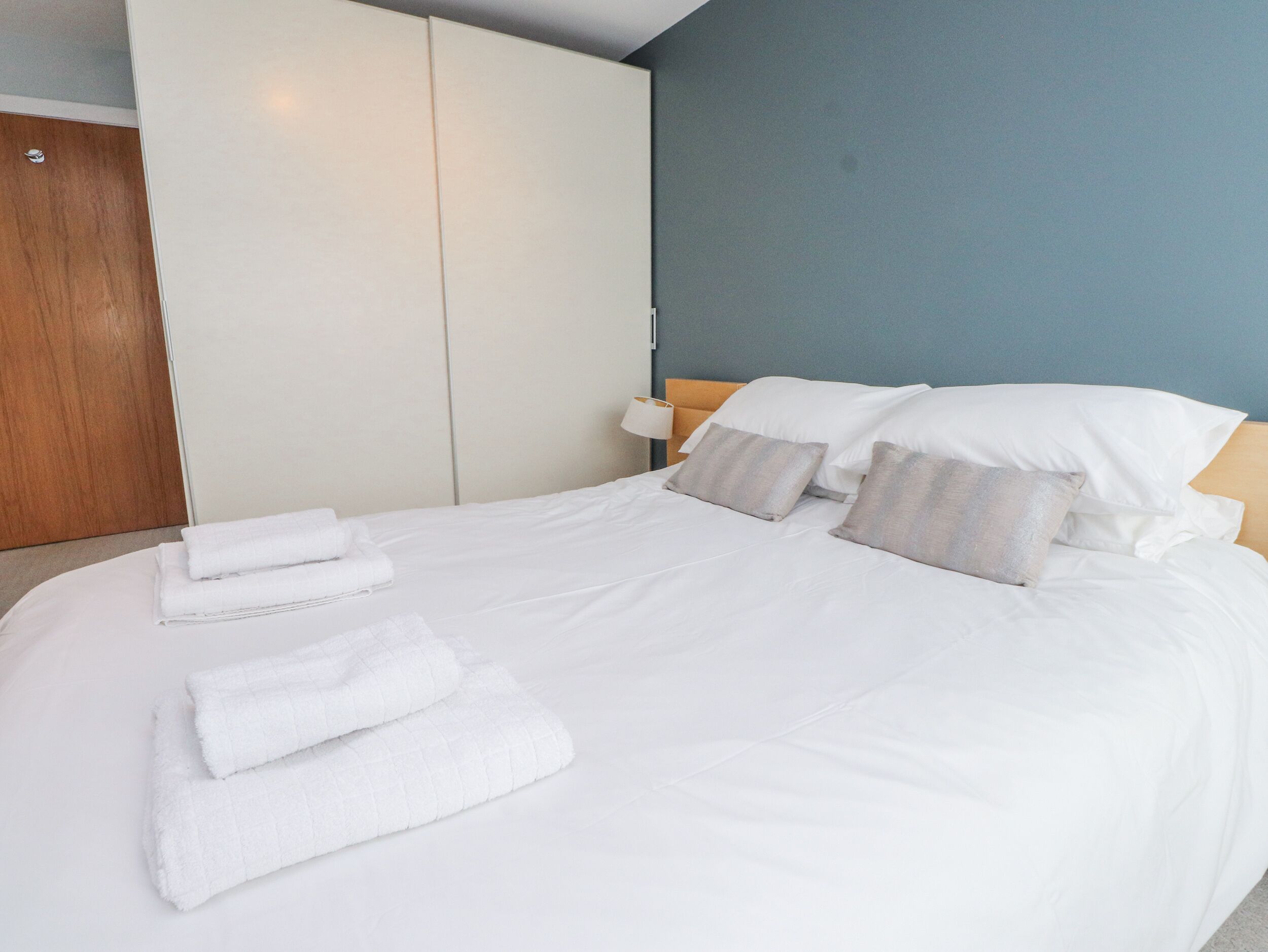 Una cama bien hecha con sábanas y almohadas blancas, un armario de madera y una pared azul.