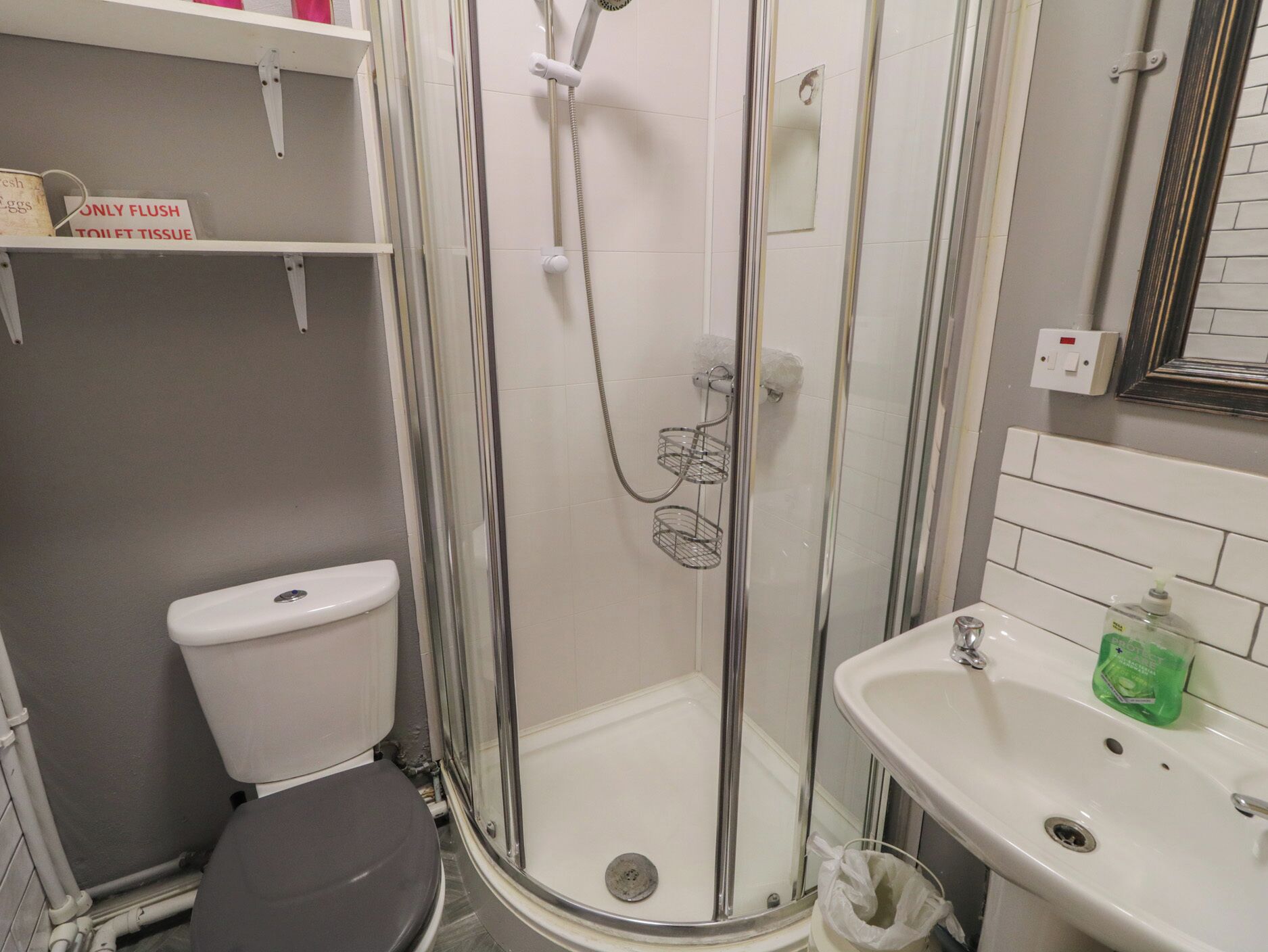A bathroom with a toilet, a shower with a glass door, a sink, and a shelf with a sign.