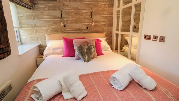 Una cama bien hecha con almohadas rosadas y grises, una manta a rayas rojas y blancas, y dos toallas enrolladas.