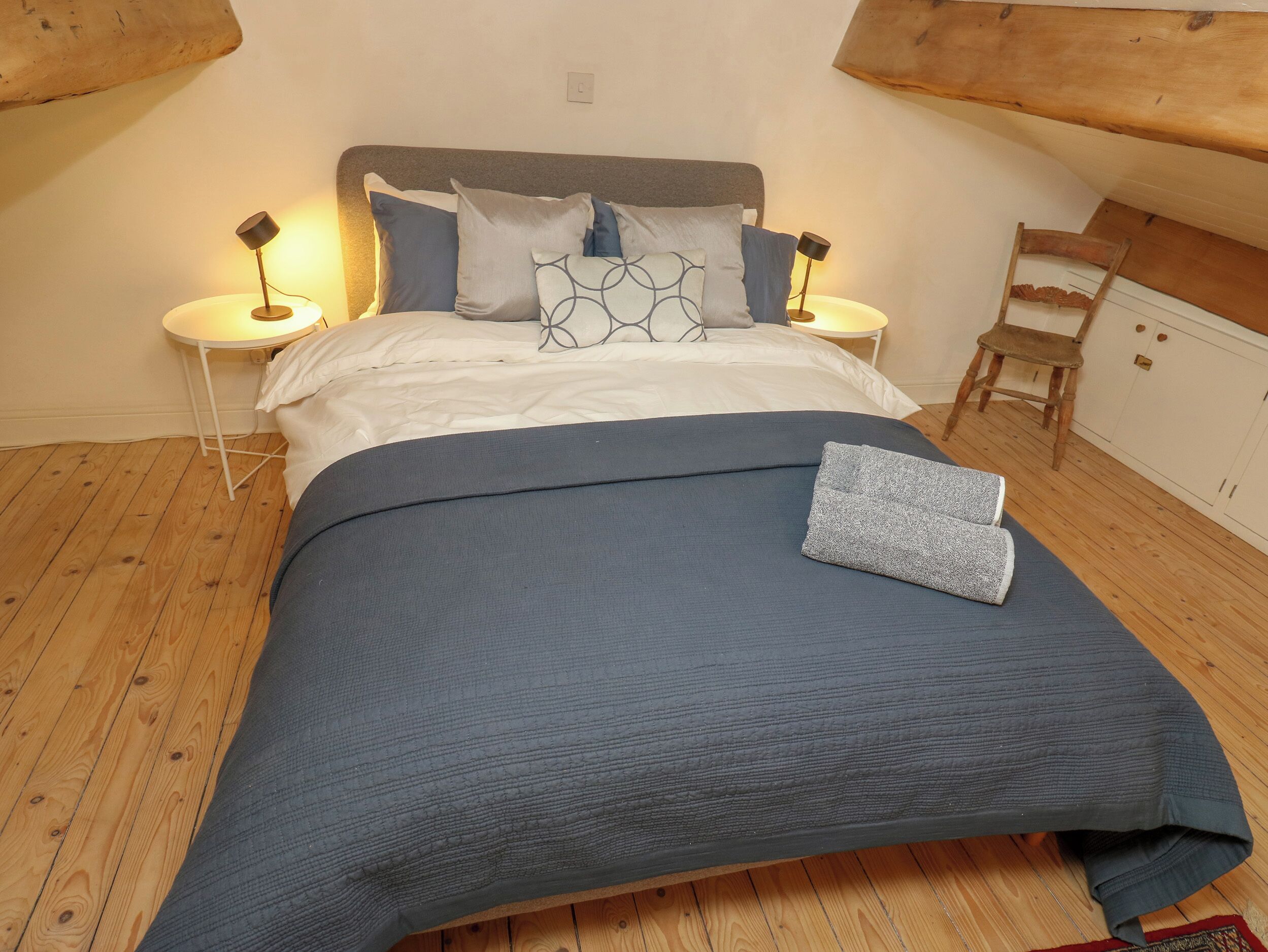 Un lit bien fait, avec une couvre-lit bleue et des oreillers, deux lampes de chevet, une chaise en bois et une armoire blanche.