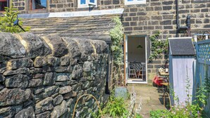 A stone house with a sloped roof, a small garden, and a wooden fence. - Cobbles Cottage (Hebden Bridge)