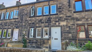 A row of terraced houses with multiple windows and doors, some with shutters, and a small garden area with plants and flowers. - Cobbles Cottage (Hebden Bridge)