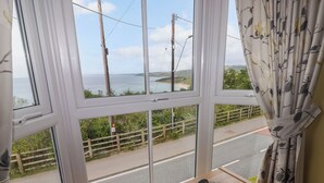 A room with a large window overlooking a road and a body of water. There is a wooden table with three spherical objects and a decorative item. - Morva (New Quay)