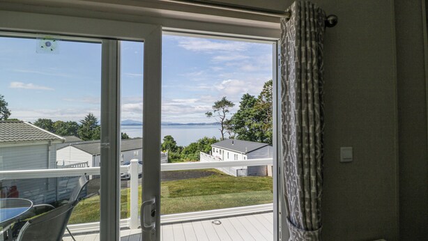 A room with a sliding glass door leading to a deck. The deck has a white railing and a glass table with chairs. There is a radiator on the wall and a curtain partially drawn to the side. - Bethany's Retreat (Wemyss Bay)
