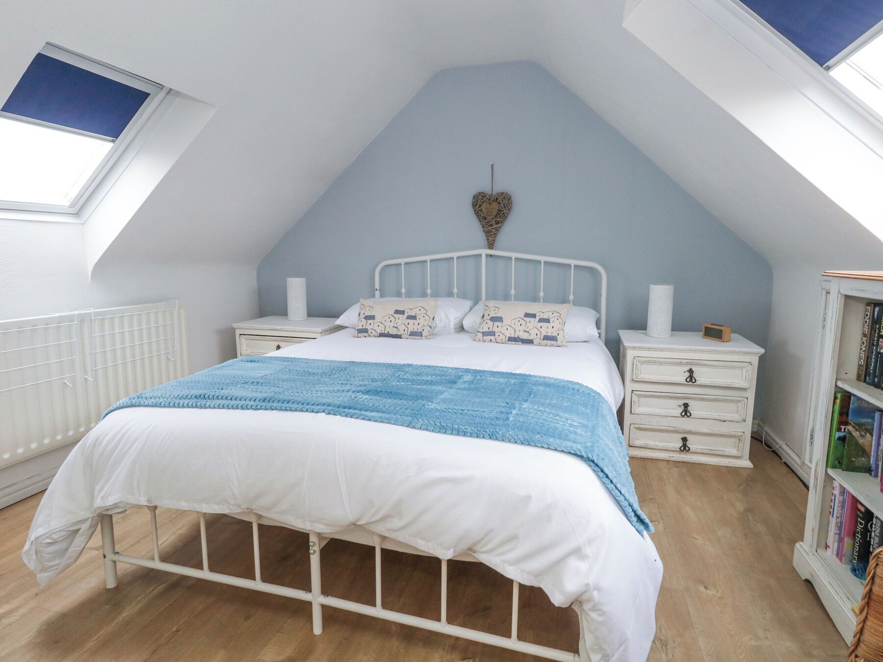 Un dormitorio con techo inclinado, una cama con ropa de cama blanca y manta azul, dos mesitas de noche con lámparas, una estantería y un tragaluz.