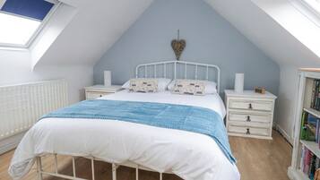 Un dormitorio con techo inclinado, una cama con ropa de cama blanca y manta azul, dos mesitas de noche con lámparas, una estantería y un tragaluz.