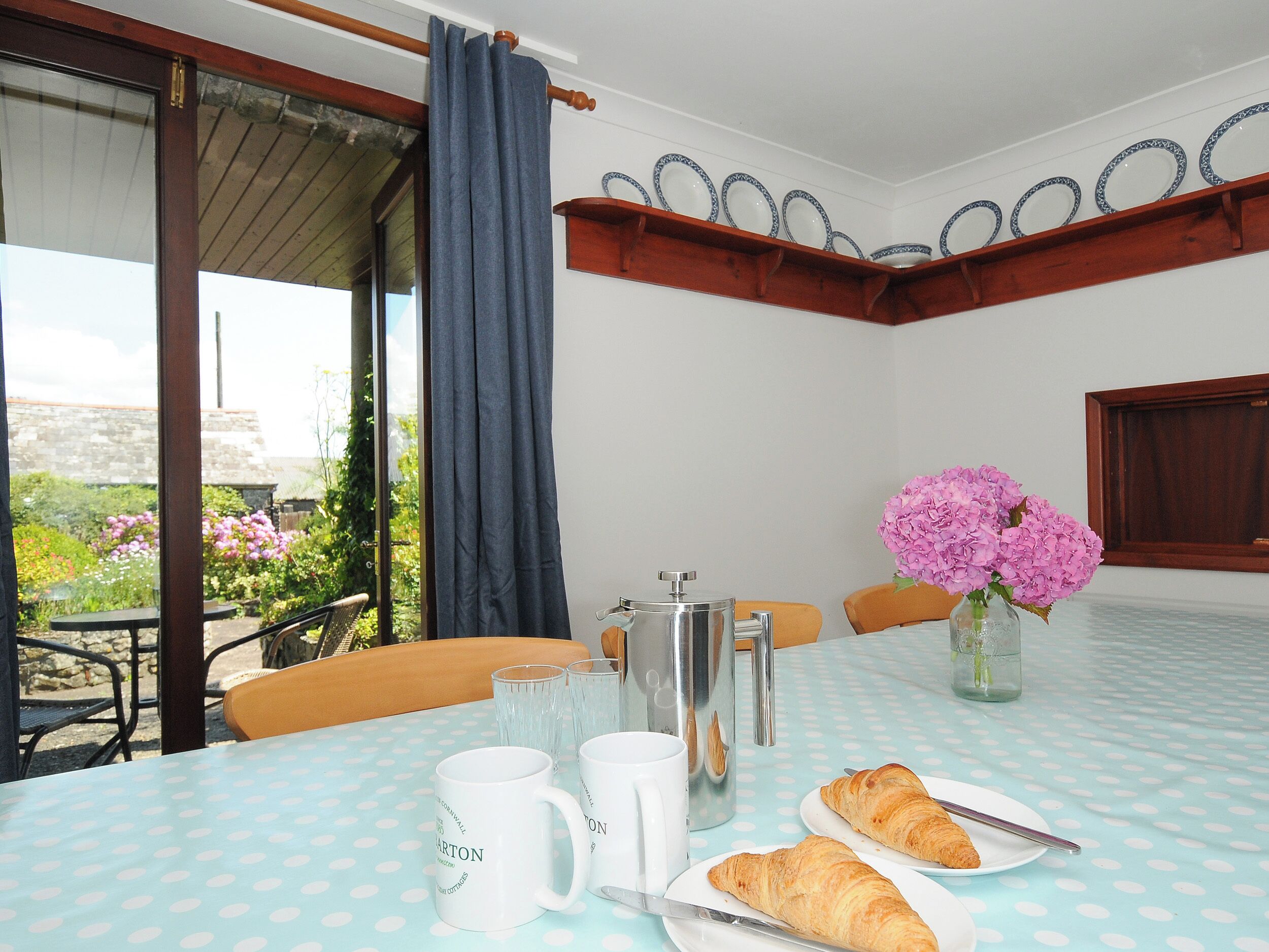 Una mesa de comedor con mantel de lunares, dos croissants, una cafetera y un florero con flores.
