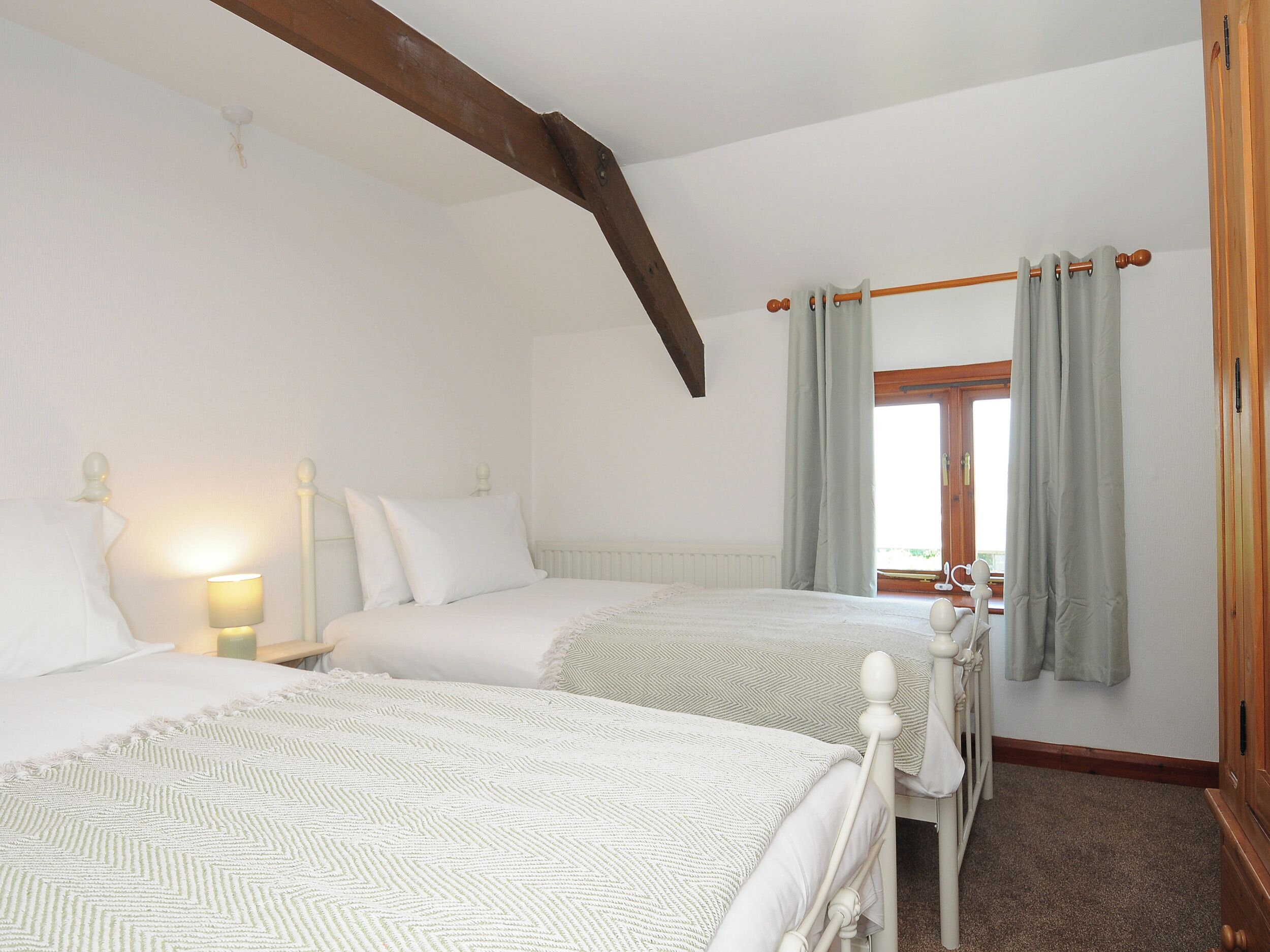 A bedroom with two beds, a wooden ceiling beam, a window with curtains, and a nightstand with a lamp.