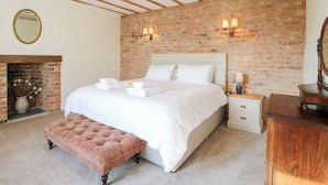A bedroom with a brick wall, wooden beams, a bed with white linens, a bench, a nightstand, a dresser, and a fireplace. - Daniels Farmhouse (Worcester)