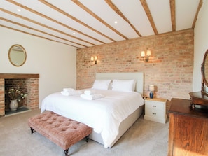 Une chambre avec un mur en briques, un plafond en bois, un lit avec du linge de lit blanc, un banc, une table de chevet, une commode et une cheminée.