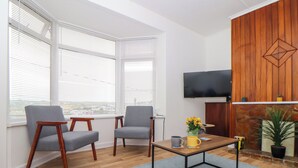 Un salon moderne avec un plancher en bois, deux fauteuils gris, une table basse, un téléviseur fixé au mur et une cheminée agrémentée d’un panneau décoratif.