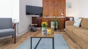 Un salon comprenant un canapé gris, un canapé marron, une table basse ornée d’un vase de fleurs, un téléviseur à écran plat fixé au mur et un mur décoratif en bois avec un motif géométrique.