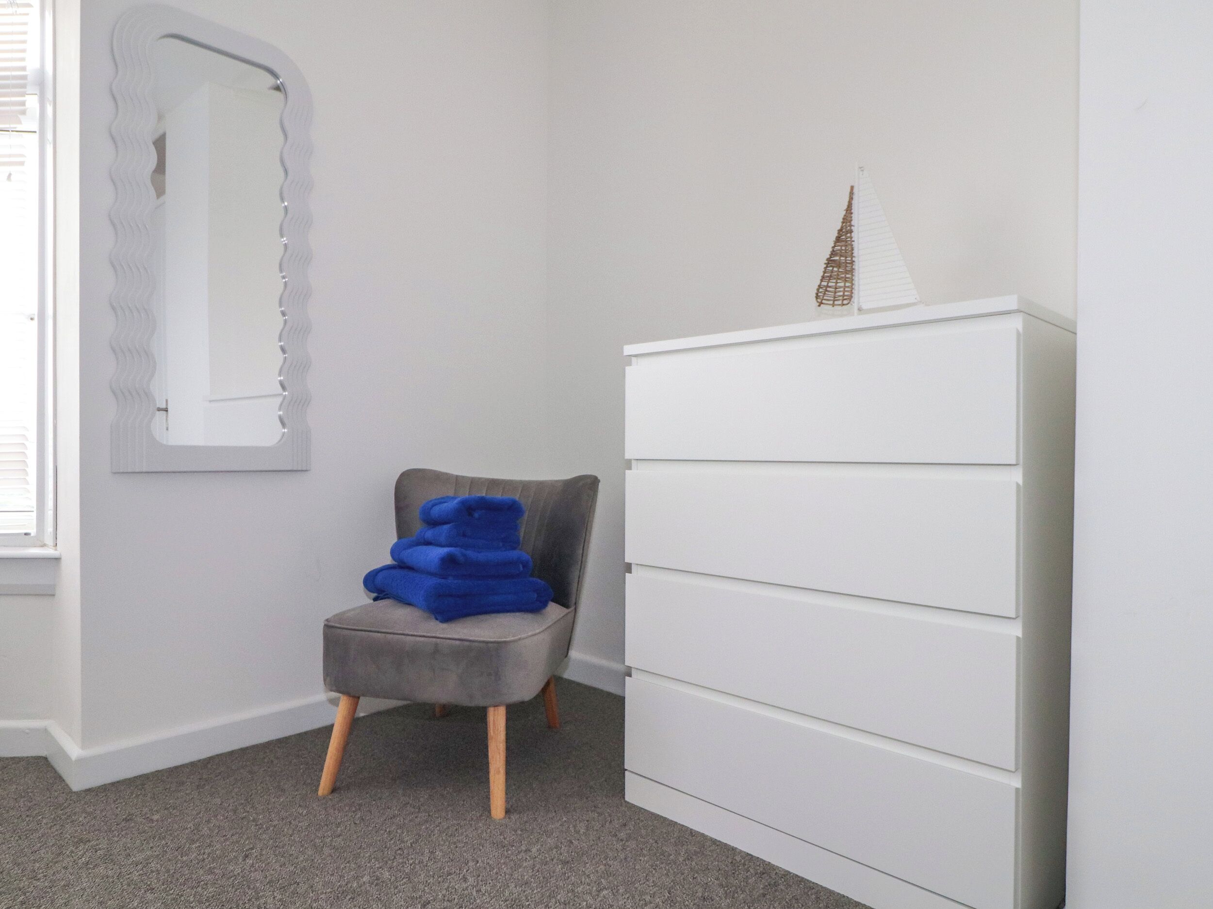 A room with a grey upholstered chair, a white dresser with multiple drawers, and a decorative item resembling a tower on top of the dresser.