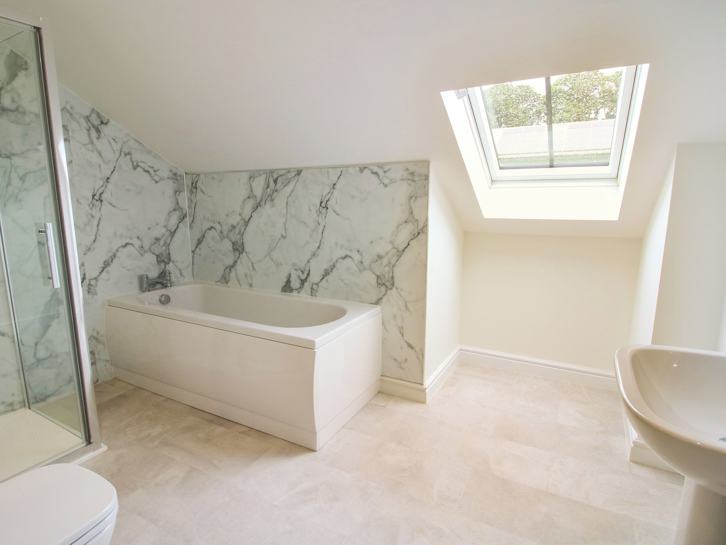 A modern bathroom with a marble wall, a freestanding bathtub, a skylight, and a separate toilet area.