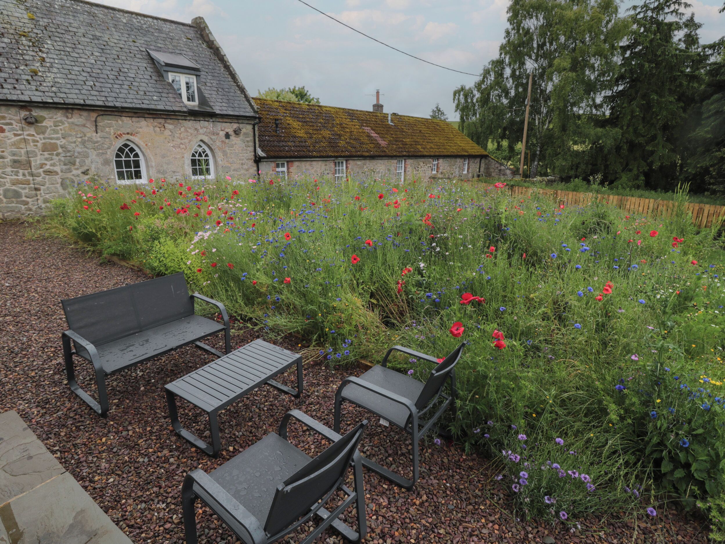 A stone building with a tiled roof, surrounded by a garden with various flowers and a gravel path.