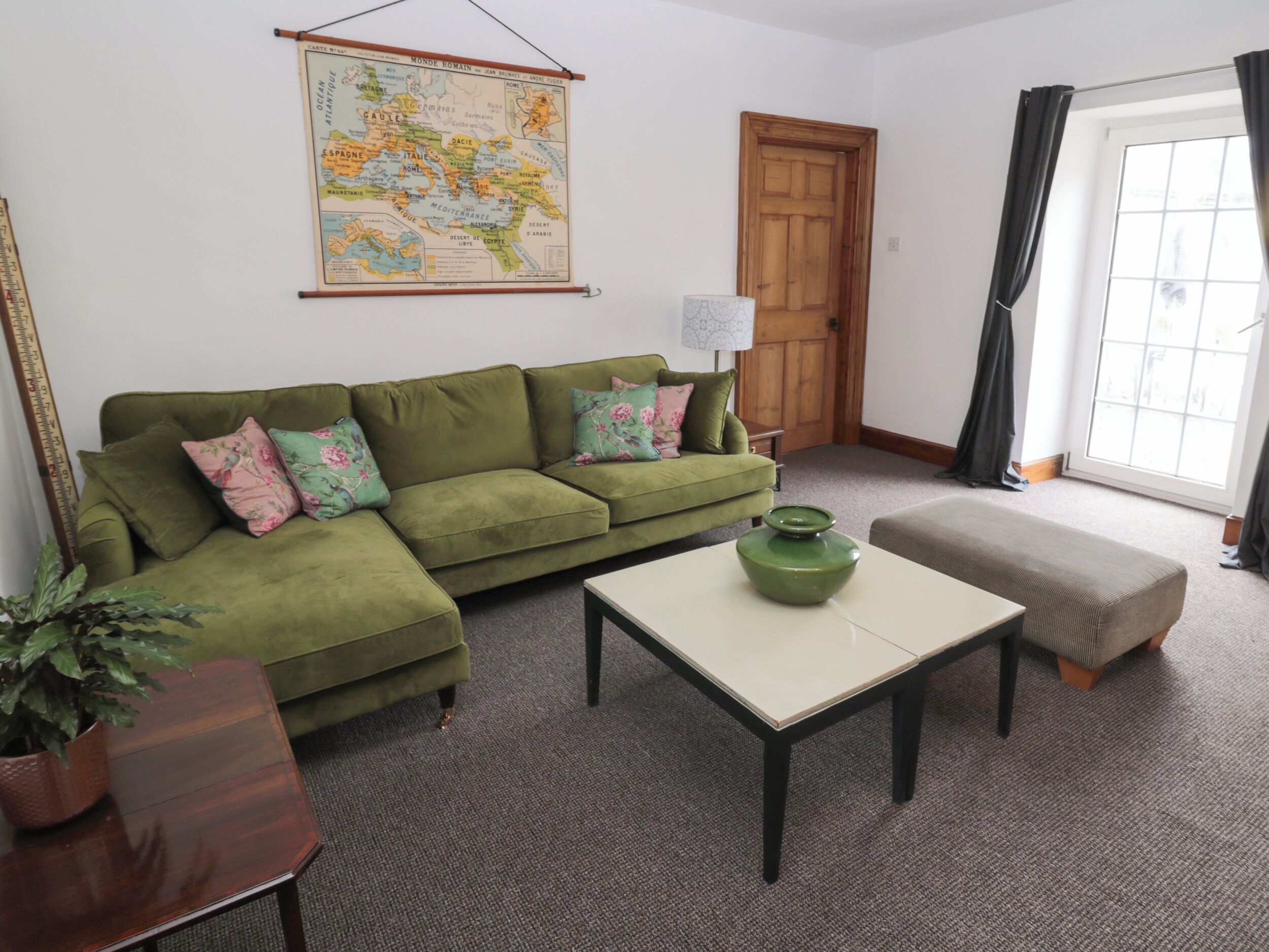 Un salon avec un canapé vert, une table basse, une carte accrochée au mur, une porte en bois et une fenêtre avec des rideaux.
