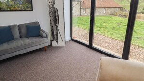 A living room with a grey sofa, a large window, a framed portrait, and a view of a house. - Honeysuckle Cottage (Cornhill on Tweed)
