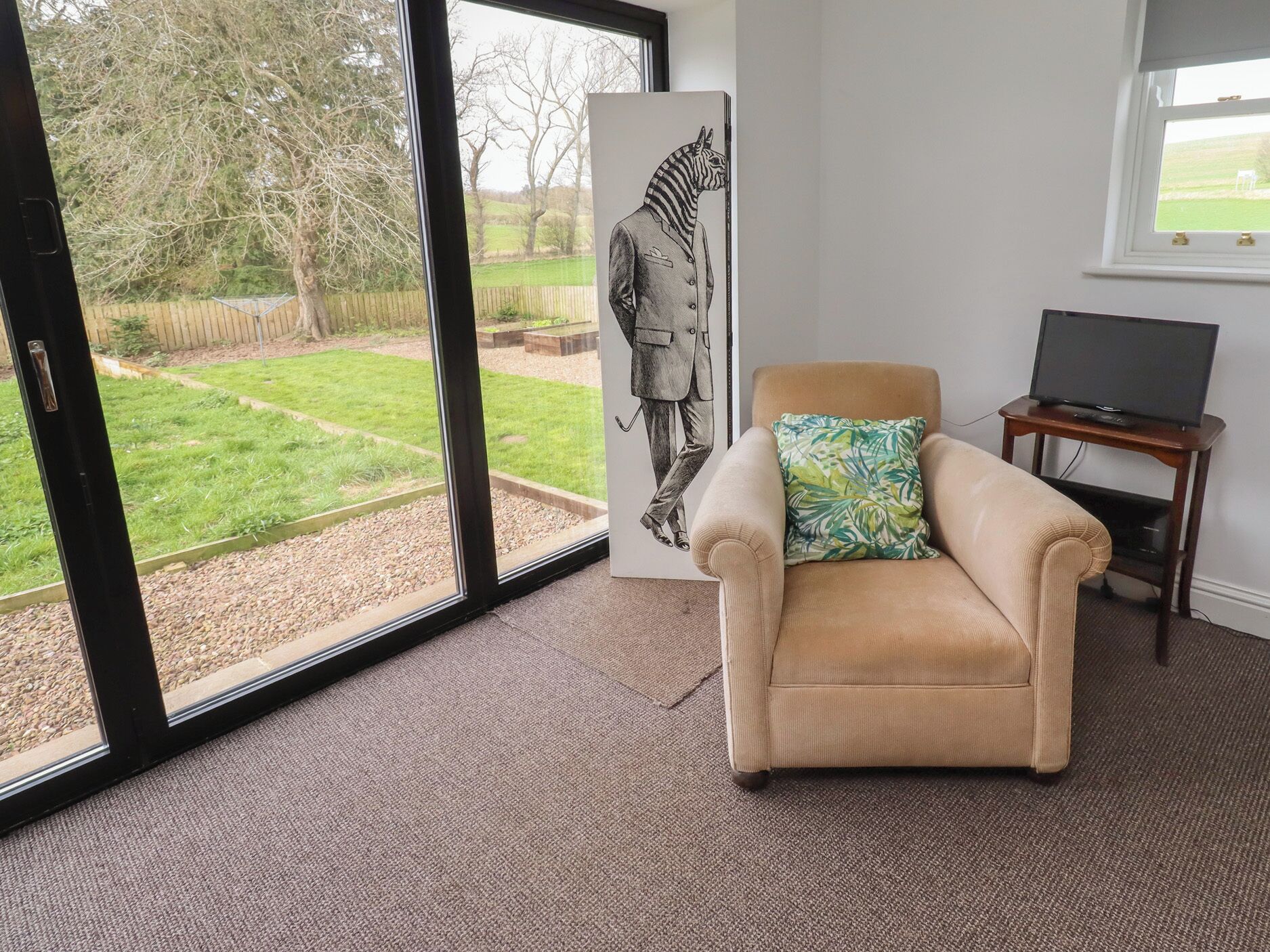 Een kamer met een groot raam, een beige fauteuil met een kussen met patroon, een televisie op een houten dressoir en een lijstwerk met een afbeelding van een zebra.
