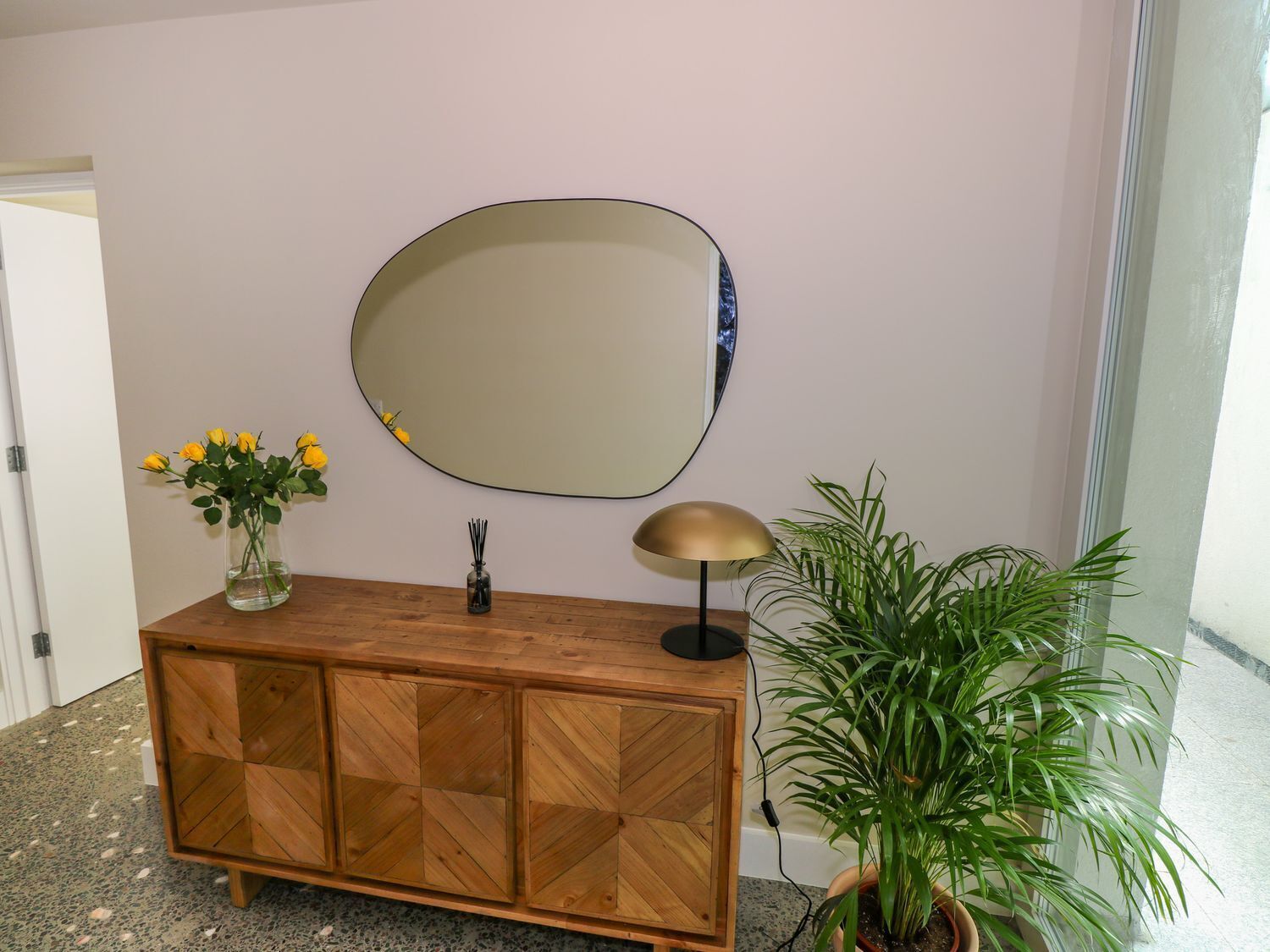 Un meuble en bois surmonté d’un miroir, un vase avec des fleurs jaunes et une plante en pot à côté.
