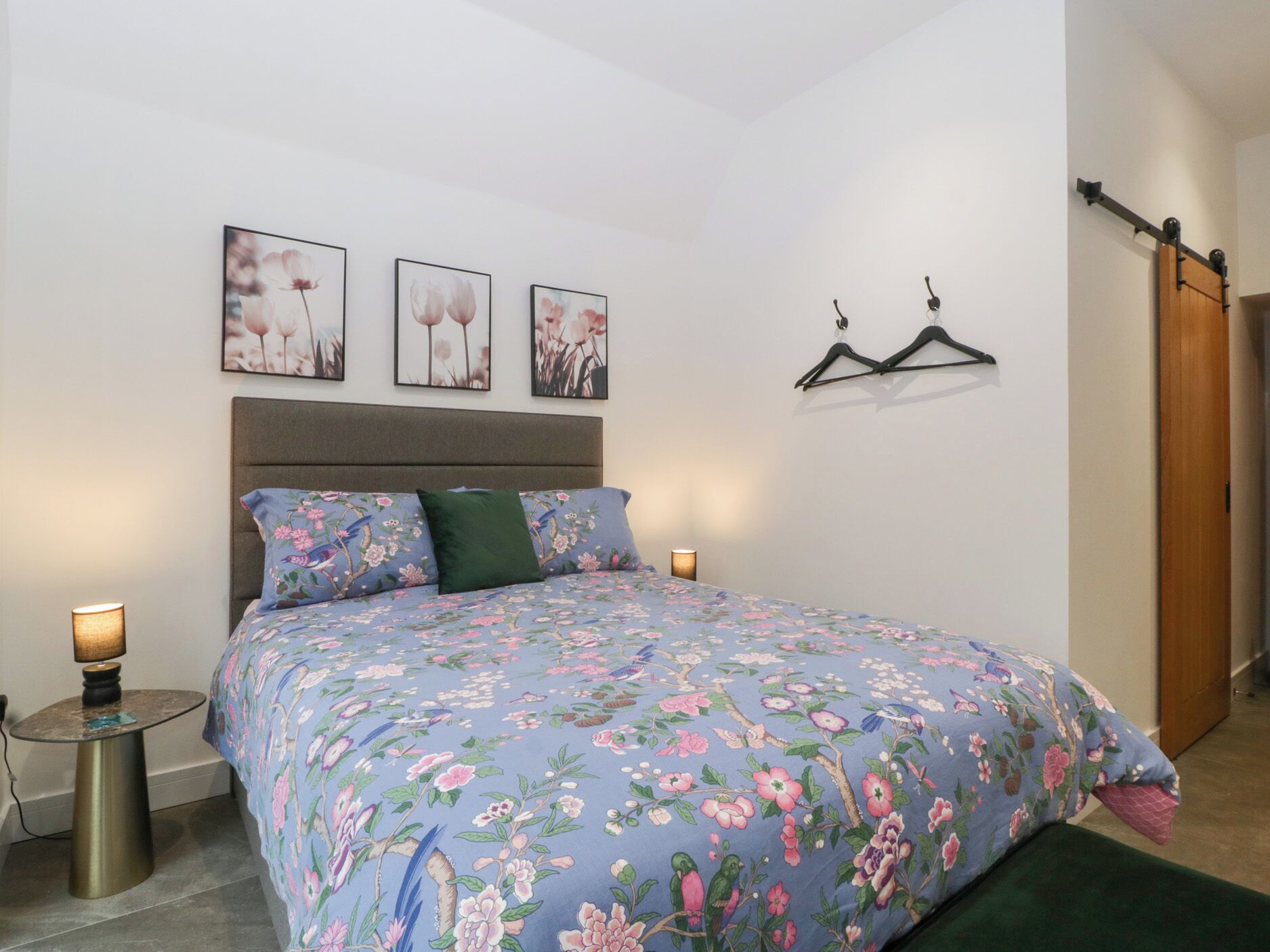 A bedroom with a floral bedspread, a bedside table with a lamp, and three wall-mounted pictures.