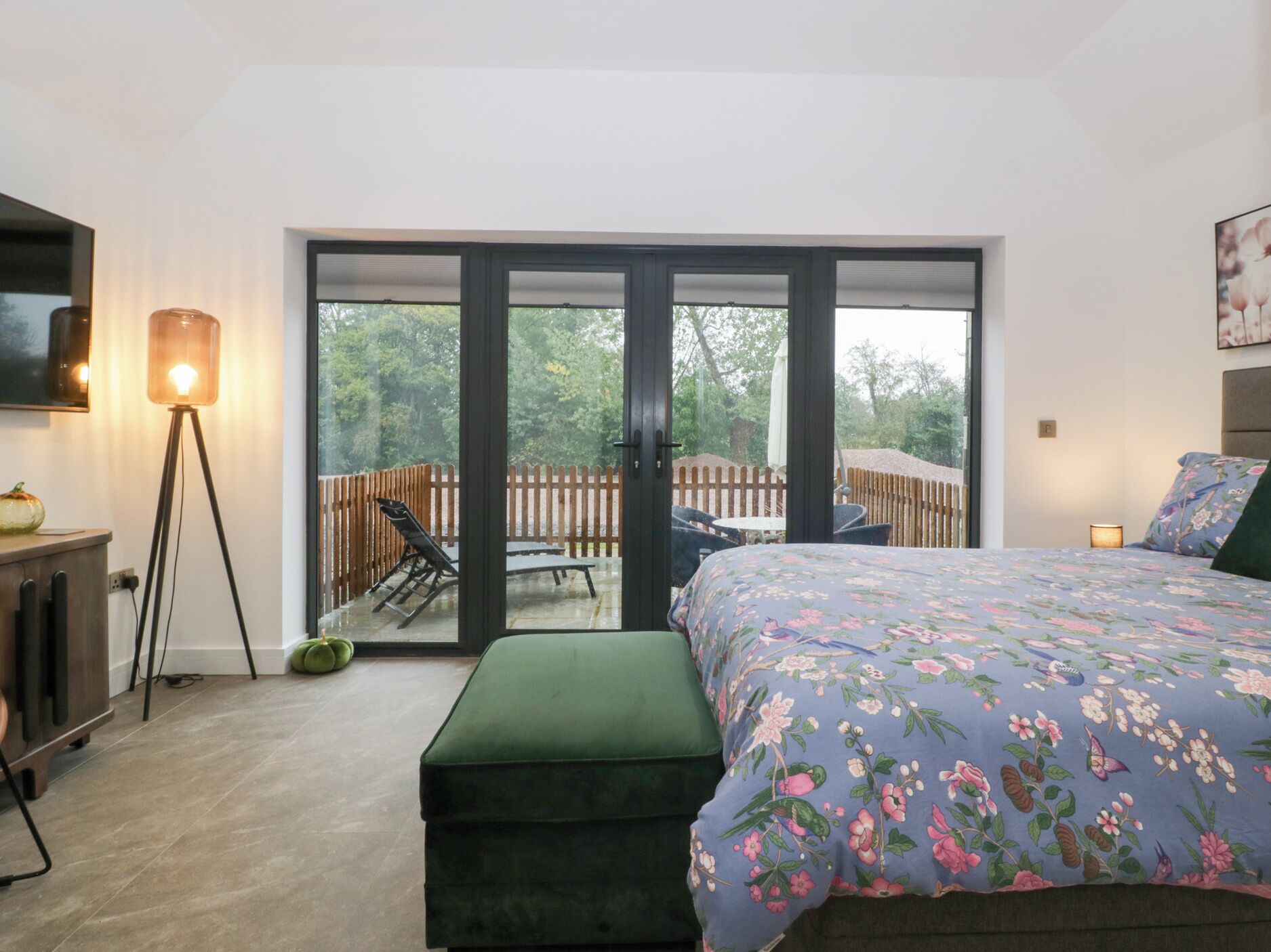 A bedroom with a bed, a green ottoman, a floor lamp, and a sliding glass door leading to an outdoor deck with a table and chairs.