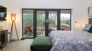 A bedroom with a bed, a green ottoman, a floor lamp, and a sliding glass door leading to an outdoor deck with a table and chairs.