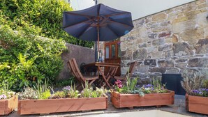 An outdoor seating area with wooden furniture and a large umbrella, surrounded by raised flower beds with various plants and flowers. - The Hayloft (St Austell)