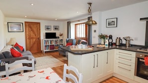A cozy living room with a sofa, a dining table with a patterned tablecloth, a kitchen area with a stove and cabinets, and a bookshelf with books and decorative items. - The Hayloft (St Austell)