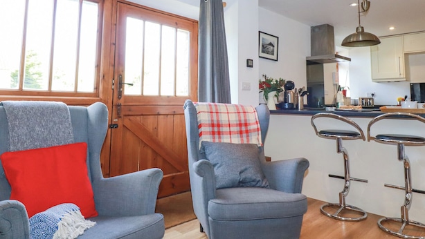 A cozy living room with two grey armchairs, a red pillow, and a blue patterned throw. A wooden door and window with curtains are visible, leading to a kitchen area with bar stools and a hanging light fixture. - The Hayloft (St Austell)