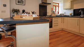A modern kitchen with a central island, wooden flooring, and a large window providing natural light. - The Hayloft (St Austell)