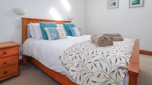 A bedroom with a wooden bed frame, white bedding, and a leaf-patterned throw. There are two wall-mounted lamps and two framed pictures on the wall. - The Hayloft (St Austell)