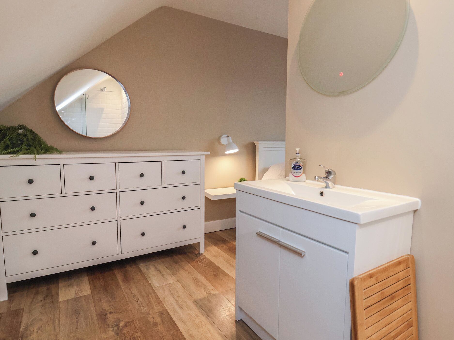 A bathroom with a white vanity, a round mirror, a sink with a soap dispenser, and a wooden bench.