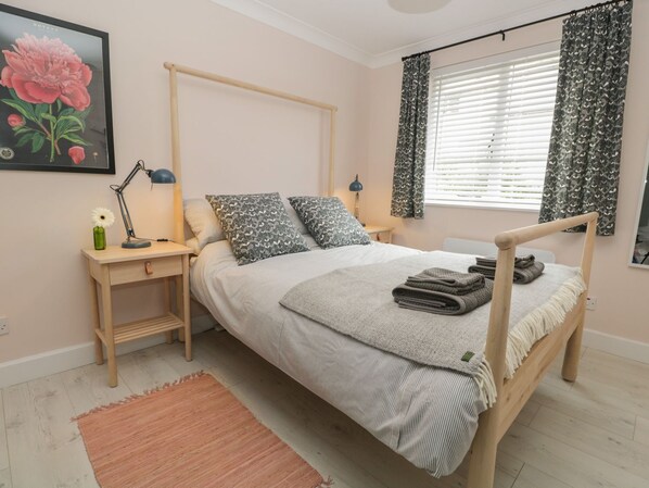 A neatly arranged bedroom with a wooden four-poster bed, two bedside tables, a nightstand, a desk, a desk lamp, a framed flower painting, a window with patterned curtains, and a small rug.