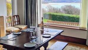 Comedor con una mesa dispuesta para cuatro personas, vista a un lago y colinas, y un ventanal con cortinas.