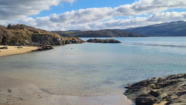 Un paisaje costero con una playa arenosa, afloramientos rocosos y un mar tranquilo bajo un cielo parcialmente nublado.