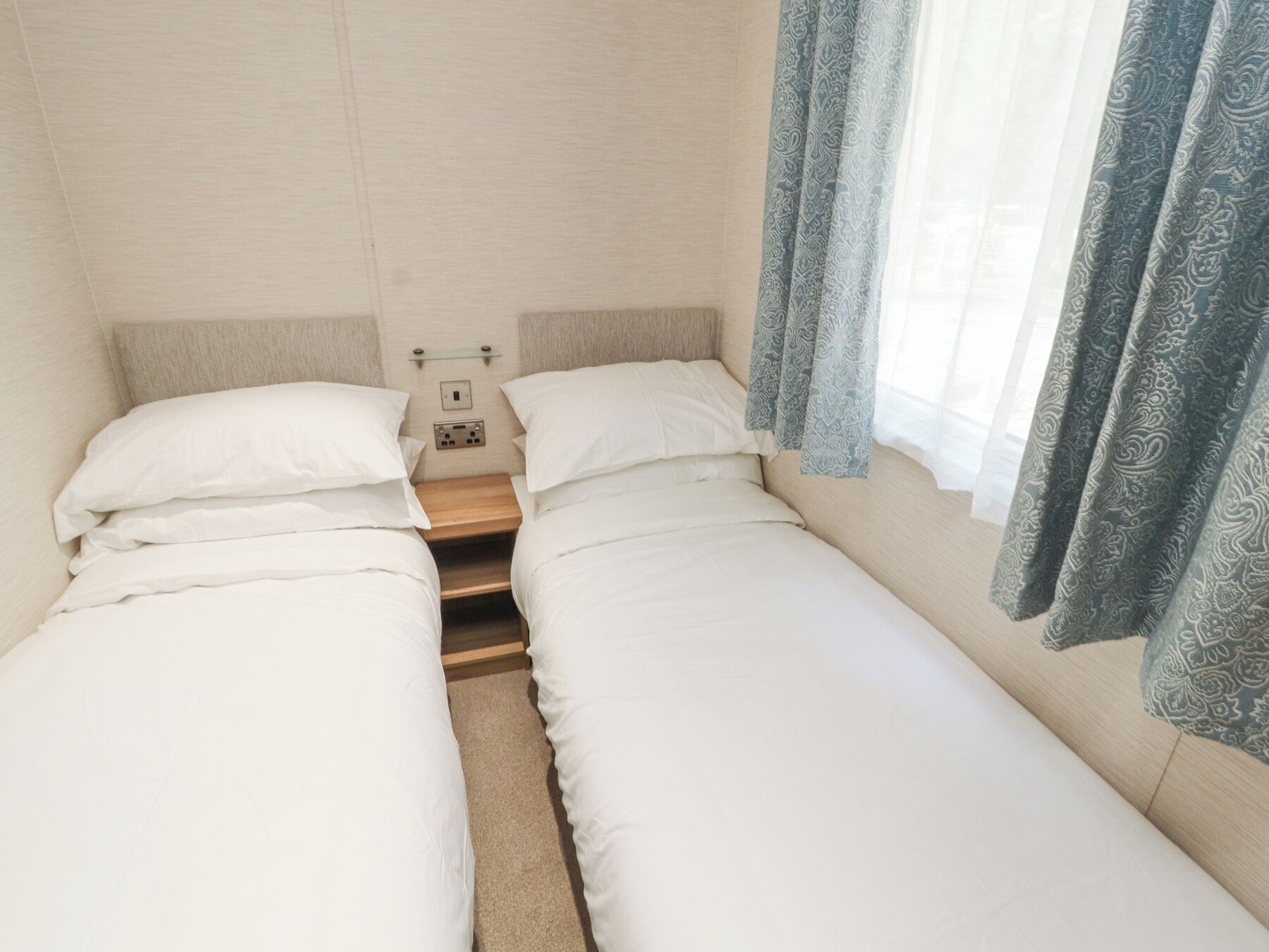 A small, clean room with two single beds, a wooden nightstand, a power outlet, and a window with patterned curtains.
