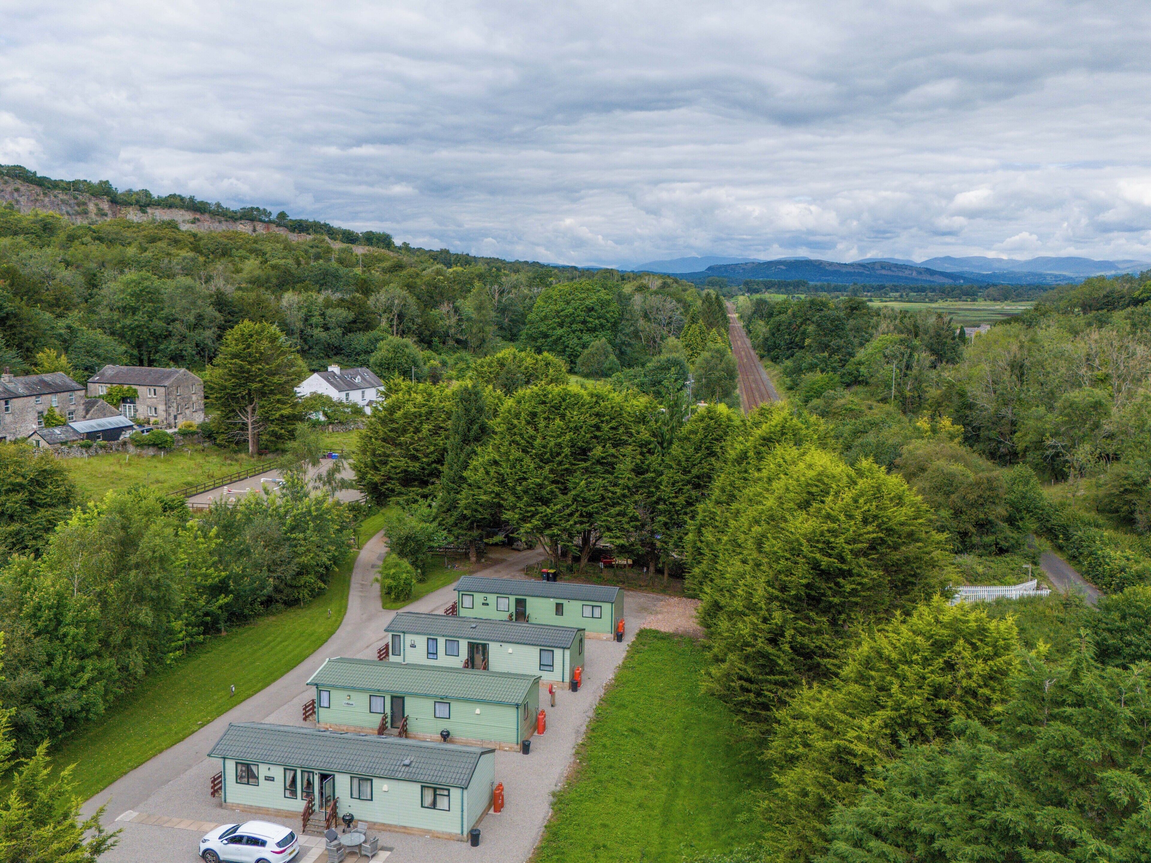 A green-roofed lodging area with multiple cabins, surrounded by lush greenery and a railway track in the background.