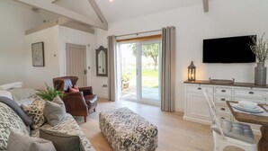 A spacious living room with a sofa, ottoman, and a dining area with a table and chairs. A television is mounted on the wall, and there is a sliding glass door leading to an outdoor area. - Church View Retreat (Weston-super-Mare)