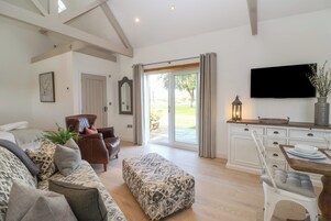 A spacious living room with a sofa, ottoman, and a dining area with a table and chairs. A television is mounted on the wall, and there is a sliding glass door leading to an outdoor area.