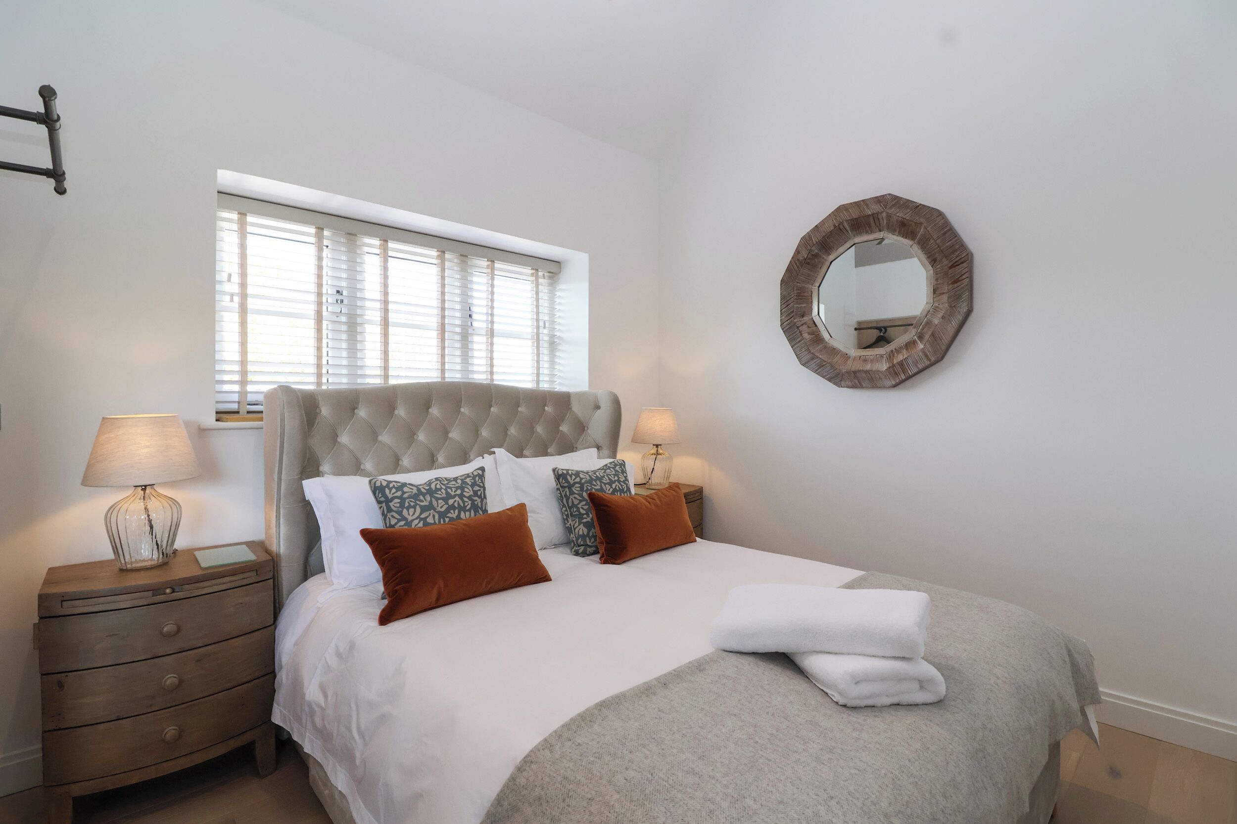 A neatly arranged bedroom with a tufted headboard, a bedside table with a lamp, and a round mirror on the wall.