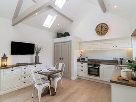 Una cocina moderna con armarios blancos, una zona de comedor con una mesa y sillas de madera, un reloj grande en la pared y un televisor sobre la encimera.