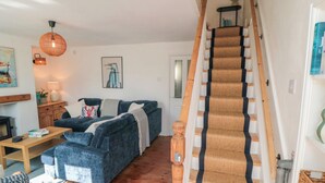 A living room with a blue sofa, wooden flooring, a staircase with a patterned carpet, and a hanging light fixture. - 6 Clarence Place (Whitby)